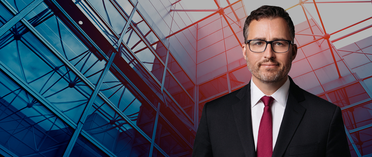 Portrait of a professional man in a dark suit and glasses, set against a modern, abstract architectural background of blue and red.